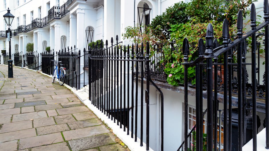 A narrow cobblestone street in South Kensington lined with residential buildings on the left, featuring white and pastel-colored facades with small windows and balconies. Potted plants and greenery de