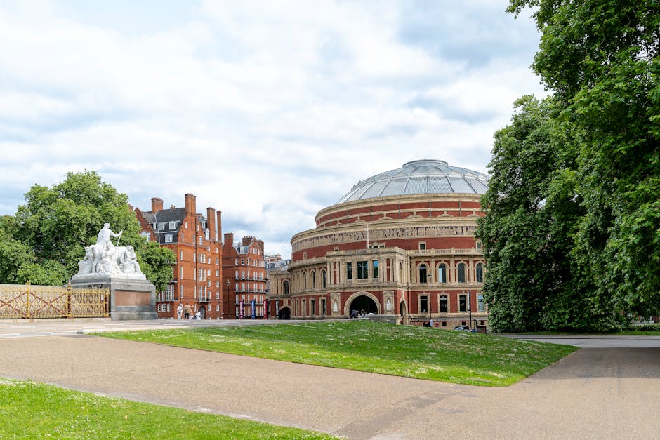 A large, historic round building with a domed roof, constructed from red brick and beige stone accents, situated in a park area with green lawns and a pathway. To the left, a white marble statue featu