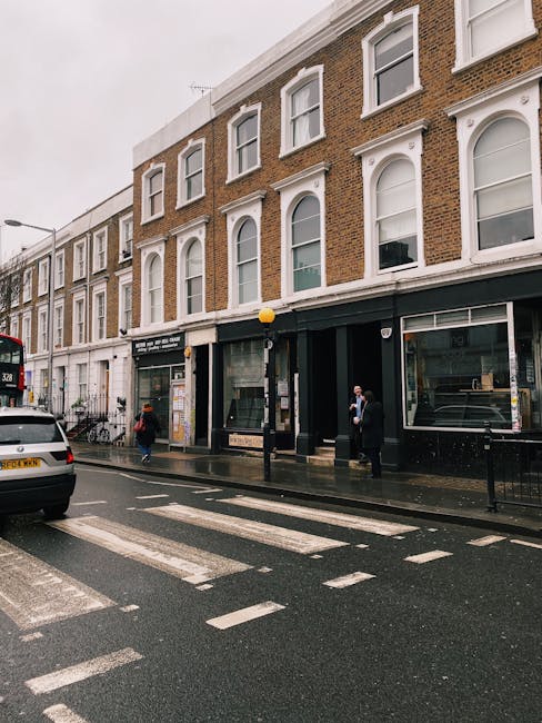 The image depicts a street scene on Gloucester Road in Kensington, featuring a row of three-story brick buildings with white window frames and decorative architectural details. The ground floors consist of commercial spaces with large glass storefronts; one appears to be a retail or service business. The street is wet, indicating recent rain, and includes a zebra crossing with white painted lines and a black lamppost. Several pedestrians, some wearing face masks, are crossing or waiting at the sidewalk. A white car and a red double-decker bus are visible on the road, and the sky appears overcast. The scene showcases urban architecture and street activity typical of Kensington, with a focus on cleanliness and well-maintained exteriors, aligned with the expertise of southkensingtoncarpetcleaners.co.uk in surface cleaning and hygiene maintenance.
