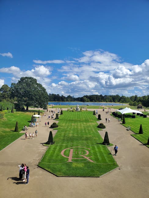 An aerial view of a large, meticulously maintained park with bright green lawns, neatly trimmed hedges, and paved walkways. The central lawn features a circular emblem with a stylized letter 'R' in pi