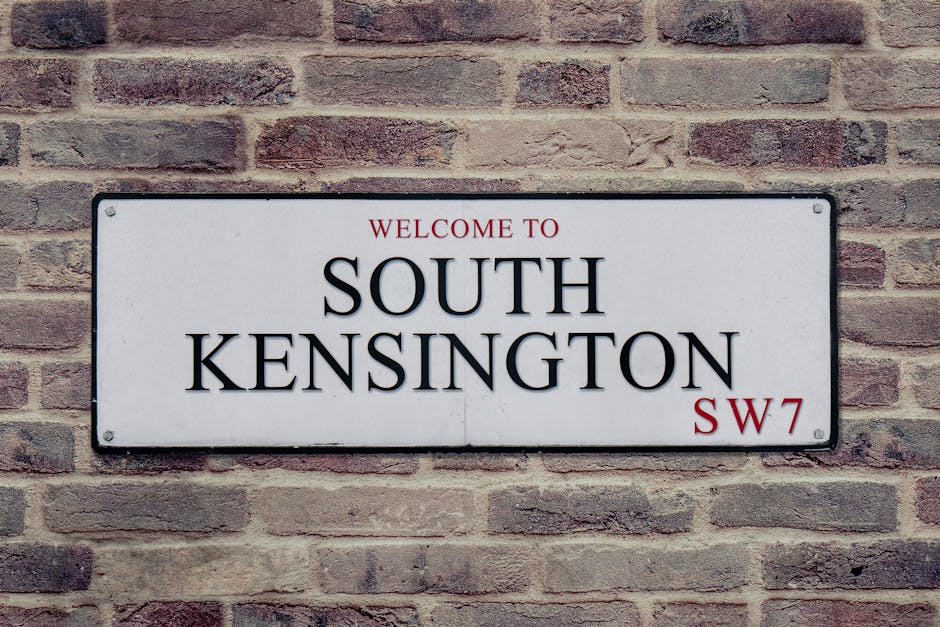 A rectangular white metal sign mounted on a brick wall displaying the text 'Welcome to South Kensington SW7' in black and red lettering. The sign is positioned centrally against a background of reddis