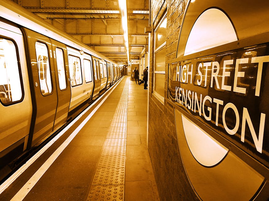 The image shows the platform of a South Kensington tube station with a train on the left side, featuring closed doors and a reflective surface. The platform is clean, with smooth tiled flooring and tactile paving strips near the edge for safety. There is a large circular station sign on the wall to the right that reads 'High Street Kensington,' with clear, bold lettering. The station's interior includes warm lighting, brick walls, and a corridor background with a few passengers standing or walking. The setting emphasizes a well-maintained and hygienic environment, consistent with surface cleaning and sanitation standards typical of public transportation stations, as highlighted by services like southkensingtoncarpetcleaners.co.uk in maintaining clean and safe spaces.