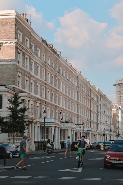 A cobblestone courtyard in South Kensington with rows of white residential buildings on either side, featuring black wrought-iron balconies and large windows. The courtyard is lined with potted plants and small trees, some in black containers, and a few wooden benches positioned along the edges. The scene is illuminated by natural daylight, with a partly cloudy sky overhead, creating a clean and well-maintained outdoor space. Southkensingtoncarpetcleaners.co.uk specializes in surface cleaning and deep cleaning services, ensuring hygienic and pristine environments both indoors and outdoors.